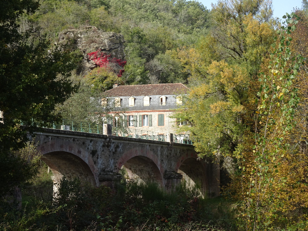Photographie du pont enjambant le Viaur et de l'auberge. Apparaissent aussi le rocher surplombant l'auberge et sa vigne vierge, ainsi que des arbres dans leurs couleurs de fin d'été.
