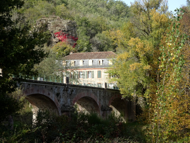 Photographie du pont enjambant le Viaur et de l'auberge. Apparaissent aussi le rocher surplombant l'auberge et sa vigne vierge, ainsi que des arbres dans leurs couleurs de fin d'été.